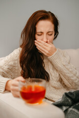 Woman coughs from a sore throat covering her face with her hand and drinks hot tea. Cold and flu concept.