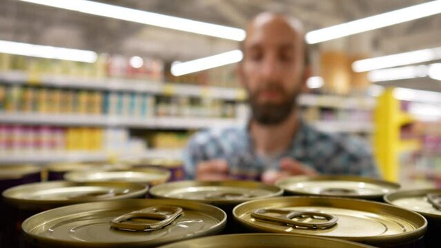 Close-up of many canned food on a supermarket shelf and a male buyer takes one