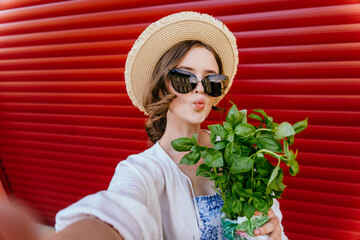 Young hipster teen girl in straw hat, sunglasses making selfie photo with bouquet of basilic with...