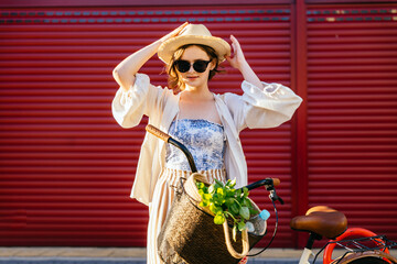 Young thoughtful woman in straw hat walking with bicycle, with reusable coffee cup in hand....