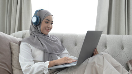 Charming young Asian Muslim woman wearing hijab sits on a comfy sofa, using a laptop computer and...