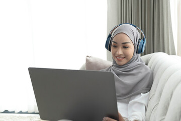 Charming young Asian Muslim woman wearing hijab sits on a comfy sofa, using a laptop computer and listening to music on her headphones.