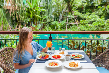 Female tourist on breakfast in restaurant at hotel. Young woman at brunch table with food and drinks. Summer holidays, vacations, tourism, travel concept. Green pond, tropical greenery background.