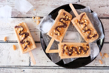 Peanut butter popsicles with chocolate drizzle on a black plate. Top view over a white wood...