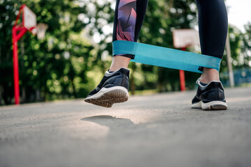 Young girl woman exercising outdoors with mini rubber elastic band doing workout. Closeup view of female person legs during active training with additional sport equipment outside - obrazy, fototapety, plakaty