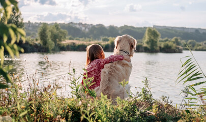 Child girl sitting and hugging golden retriever dog outdoors at the nature and looking at the lake....