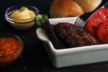 Cutlets with vegetables in a white plate with containers of sauces under dark lighting. Shallow depth of field