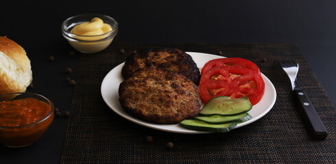 Cutlets with vegetables in a white plate with containers of sauces under dark lighting. Shallow depth of field