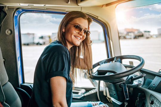 Portrait Of Beautiful Young Woman Professional Truck Driver Sitting In A Big Truck, Looking At Camera And Smiling. Inside Of Vehicle. People And Transportation Concept.