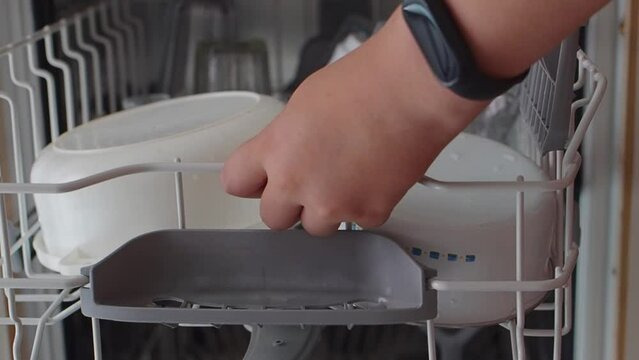 Teenager boy puts dirty dishes in the dishwasher. Young guy hands unload the dishwasher in close-up. Modern household appliances at home. Young guy puts the top baskets with dirty dishes in dishwasher