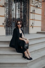 a brunette girl in a black jacket and dress and black glasses sits in the city with a handbag