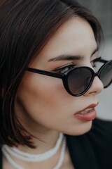 a brunette girl in a black jacket and dress and black glasses sits in the city with a handbag