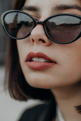 a brunette girl in a black jacket and dress and black glasses sits in the city with a handbag