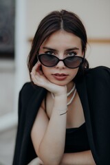 a brunette girl in a black jacket and dress and black glasses sits in the city with a handbag