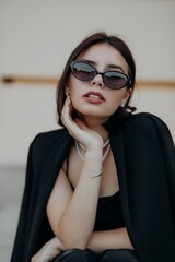 a brunette girl in a black jacket and dress and black glasses sits in the city with a handbag