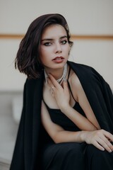 a brunette girl in a black jacket and dress and black glasses sits in the city with a handbag