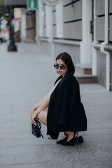 a brunette girl in a black jacket and dress and black glasses sits in the city with a handbag
