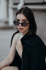 a brunette girl in a black jacket and dress and black glasses sits in the city with a handbag