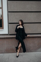 a brunette girl in a black jacket and dress and black glasses walks down the street in the city with a handbag