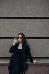 a brunette girl in a black jacket and dress and black glasses walks down the street in the city with a handbag