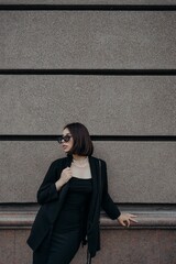 a brunette girl in a black jacket and dress and black glasses walks down the street in the city with a handbag