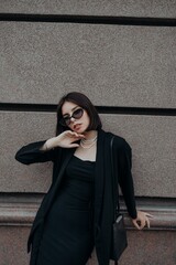 a brunette girl in a black jacket and dress and black glasses walks down the street in the city with a handbag