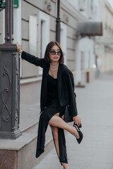 a brunette girl in a black jacket and dress and black glasses walks down the street in the city with a handbag