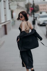 a brunette girl in a black jacket and dress and black glasses walks down the street in the city with a handbag