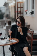 a brunette girl in a black jacket and dress and black glasses sits in a cafe with a handbag and a magazine and paints her lips