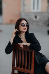 a brunette girl in a black jacket and dress and black glasses sits in a cafe