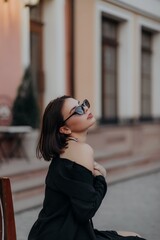 a brunette girl in a black jacket and dress and black glasses sits in a cafe
