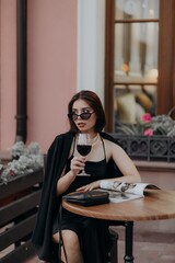 a brunette girl in a black jacket and dress and black glasses sits in a cafe with a handbag and a magazine and drinks wine