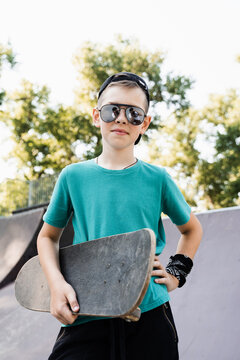 Fashion child boy in glasses posing with skate board on sport playground. Active teenager with sport equipment on sport ramp. Extreme lifestyle.