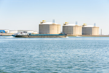 Bulk carrier barge in harbour with large concrete tanks for storage of liquefied natuaral gas in background. Port of Rotterdam, Netherlands. - obrazy, fototapety, plakaty