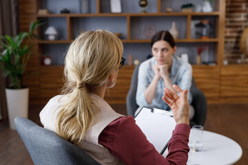 Over shoulder view of female psychologist sitting in armchair, talking with upset woman patient....