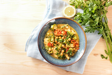 Indian style curry from chickpeas, colorful vegetables and chicken meat in a blue bowl, lemon, herbs and napkin on a light wooden table, copy space, high angle view