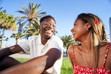 Obraz premium Cheerful young African couple laughing and having fun on their first date in the park in a romantic outdoor lovers scene, sitting on the grass talking