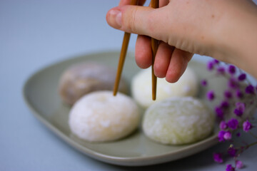 Chopsticks in a hand of a person going to eat a traditional Japanese dessert mochi in rice dough or daifuku. Mochi ice cream balls on gray plate on blue background. Purple flowers decorating a dish.