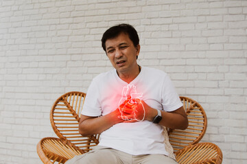 Middle aged, senior Asian man with sudden onset of heart disease showing pain heart muscle leaks tired and suffocating sits on a wooden chair in agony : Myocardial infarction (Heart Attack) - obrazy, fototapety, plakaty