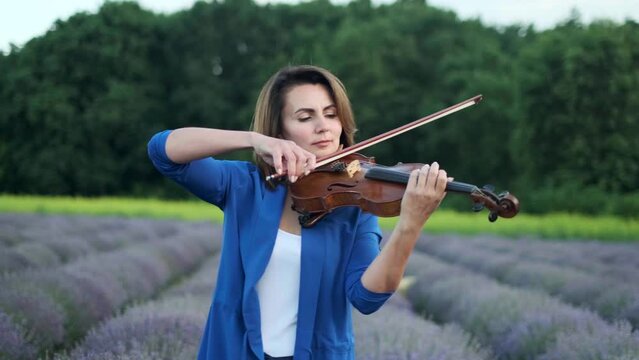 Adult woman violinist playing violin on summer lavender field in evening, romantic musician in blue dress enjoying walking on nature