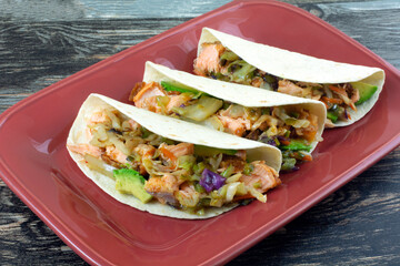 Three salmon tacos with avocado and vegetables on red serving plate