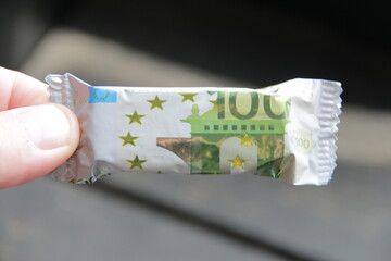 A hand holds a candy with a wrapper in the form of a one hundred euro banknote