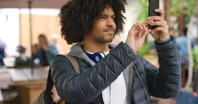 Happy student on campus taking pictures with his phone. Young man on university or college campus with wireless headphones around his neck taking photos of his educational facility while learning