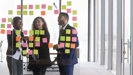 Happy diverse millennial business team meeting at board with paper sticky notes, discussing project tasks, business process, strategy, schedule, reading memos, smiling, laughing. Through glass view