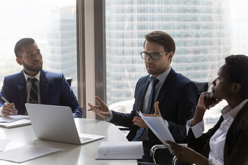 Confident Caucasian businessman, team leader speaking at corporate meeting. Diverse colleagues, business partners, discussing project, brainstorming, negotiating on deal in office