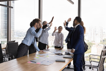Happy excited business colleagues giving group high five over meeting table with reports, celebrating team success, achieve, good job result, profit, successful project startup. Teamwork concept