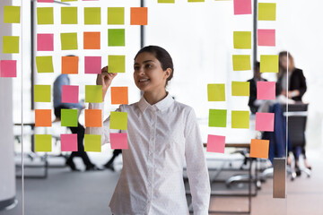 Happy confident young Indian business professional making project schedule, writing on paper sticky...
