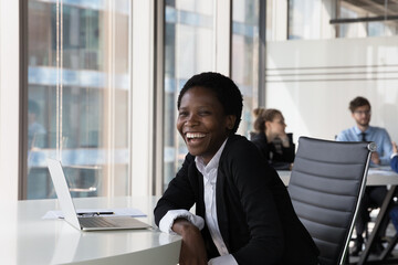 Happy excited African employee woman laughing at computer at office workplace table, looking at camera. Successful confident young business woman getting good news, enjoying career success