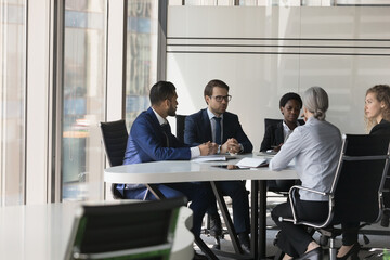 Serious diverse group of business coworkers collaborating on project. Business partners negotiating on deal, discussing agreement terms at meeting table in modern office space interior
