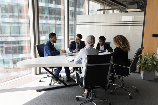 Millennial diverse different aged business team meeting in modern office space. Partners, stakeholders, colleagues negotiating at conference table at panoramic glass wall window